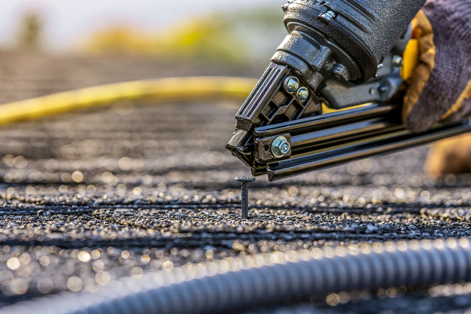 A pneumatic nail gun in a roofer's hand driving a shingle nail