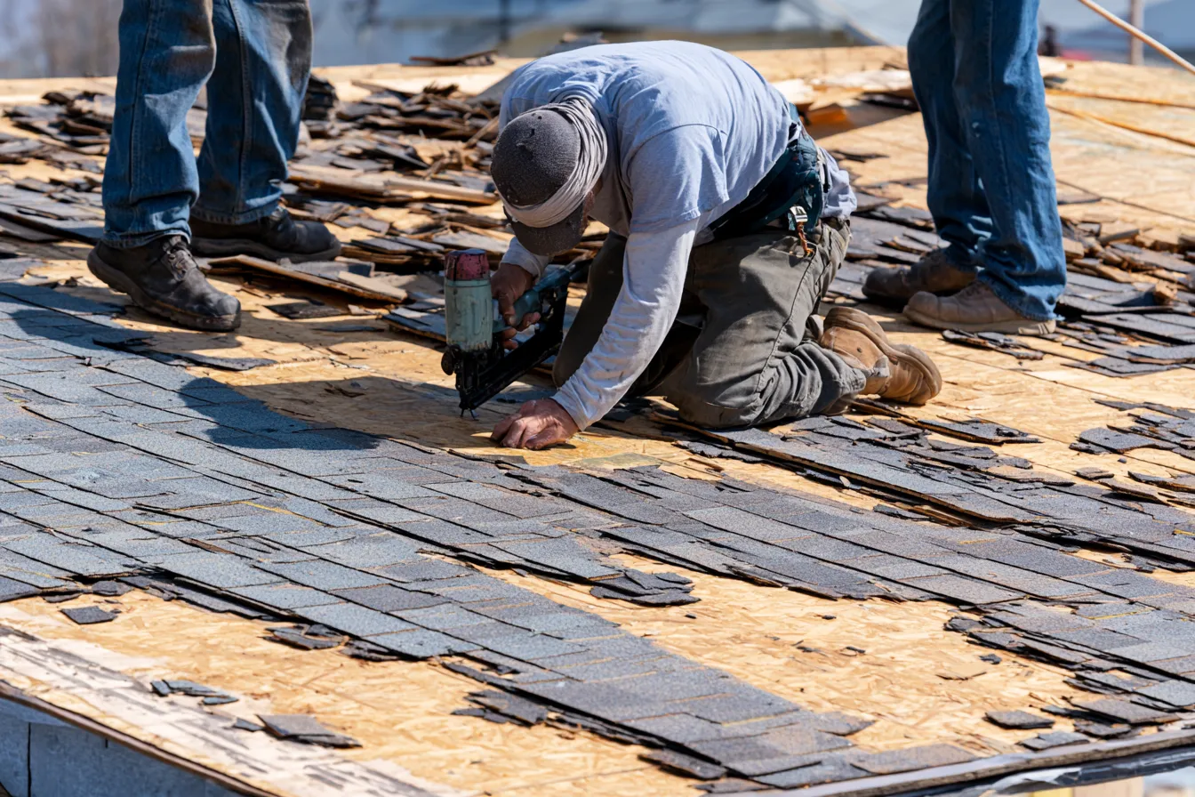 Nail gun held by a kneeling roofer on partially stripped roof decking